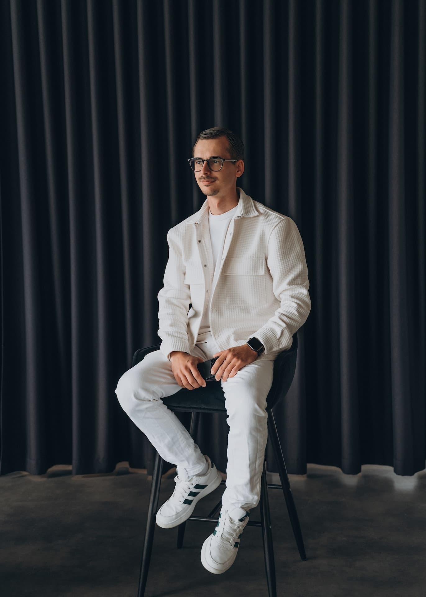 Vigo Krumins in white, seated on a bar stool in front of a dark curtain